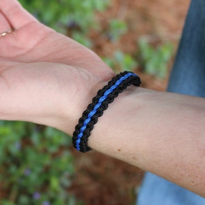 May include: Black and blue braided bracelet. The bracelet features a central blue stripe between two black braided sections. The bracelet is worn on a wrist, with a blurred background of greenery and a hand holding a pink flower.