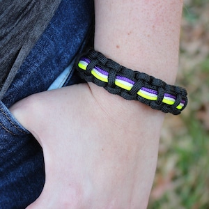 May include: Black paracord bracelet with interwoven neon yellow, white, and purple strands. The bracelet is worn on a wrist, with a hand in a blue jeans pocket. The bracelet is a handmade accessory.