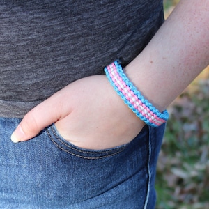 May include: A woven bracelet in shades of light blue, pink, and white. The bracelet is worn on a wrist, with the person's hand in a blue jeans pocket. The bracelet is approximately 0.75 inches wide.