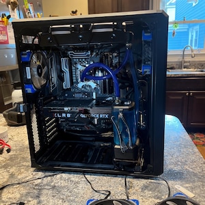 May include: A black computer case with internal components visible, including a graphics card labeled "GeForce RTX" and a liquid cooling system with blue tubing. Two Corsair fans with blue accents are in the foreground.