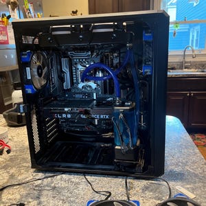 May include: A black computer case with internal components visible, including a graphics card labeled "GeForce RTX" and a liquid cooling system with blue tubing. Two Corsair fans with blue accents are in the foreground.