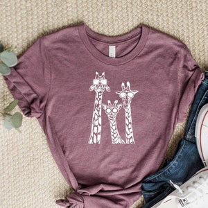 May include: A maroon t-shirt featuring a white graphic of three giraffes wearing sunglasses. The giraffes are standing side by side, with the tallest giraffe at the back and the shortest giraffe at the front.