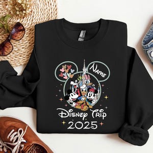 Op de afbeelding: Zwart sweatshirt met een Mickey Mouse-ontwerp en de tekst "Disney Trip 2025". Het ontwerp bevat Mickey Mouse, Minnie Mouse, Donald Duck, Goofy, Pluto en andere Disney-figuren. Het sweatshirt heeft een ronde hals en lange mouwen.
