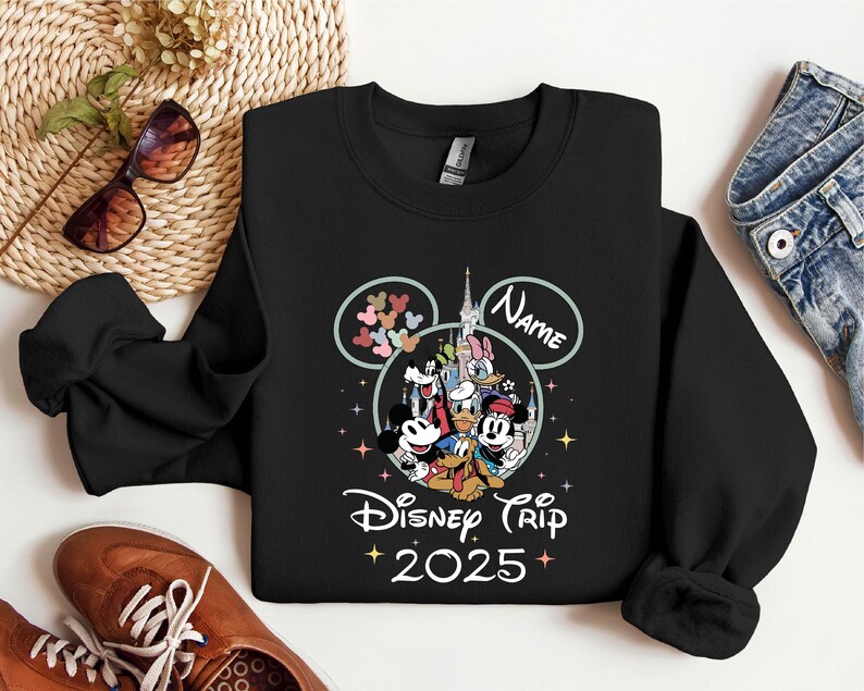 Op de afbeelding: Zwart sweatshirt met een Mickey Mouse-ontwerp en de tekst "Disney Trip 2025". Het ontwerp bevat Mickey Mouse, Minnie Mouse, Donald Duck, Goofy, Pluto en andere Disney-figuren. Het sweatshirt heeft een ronde hals en lange mouwen.
