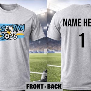 May include: A light gray t-shirt with the text "ARGENTINA 2026" in blue and yellow, featuring a soccer ball graphic. The back of the shirt has "NAME HERE" and the number 1 in black. A soccer ball is visible in the foreground.