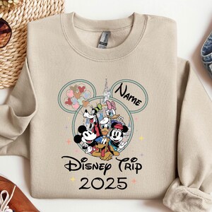 Op de afbeelding: Een beige sweatshirt met een Mickey Mouse-kopontwerp. Het ontwerp toont Mickey Mouse, Minnie Mouse, Donald Duck, Goofy, Pluto en een kasteel. De tekst "Disney Trip 2025" staat onder het ontwerp. De tekst "Name" staat in een cirkel boven het ontwerp.