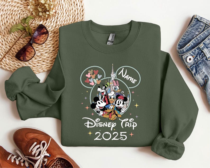 Op de afbeelding: Een groen sweatshirt met een Mickey Mouse en vrienden ontwerp. Het ontwerp bevat Mickey Mouse, Minnie Mouse, Donald Duck, Goofy, Pluto en een kasteel. De tekst "Disney Trip 2025" is onder het ontwerp gedrukt. De tekst "Name" is boven het ontwerp gedrukt.