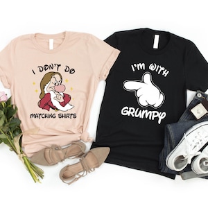 Könnte beinhalten: Zwei T-Shirts, eines hellrosa und eines schwarz, mit einem passenden Partner-Design. Das rosa Shirt zeigt ein Cartoon-Bild von Doc aus Schneewittchen und den Text "Ich mag keine passenden Shirts". Das schwarze Shirt zeigt eine weiße Cartoon-Hand, die zeigt, und den Text "Ich bin mit Grumpy".