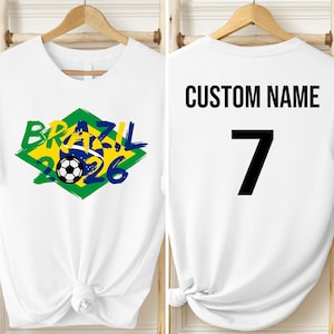 May include: White t-shirt with a Brazil 2026 soccer graphic in green, yellow, and blue. The front design includes a soccer ball. The back of the shirt has the text "CUSTOM NAME" above the number 7 in black.
