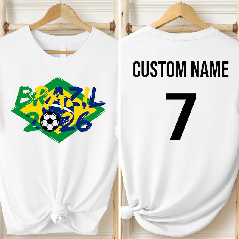 May include: White t-shirt with a Brazil 2026 soccer graphic in green, yellow, and blue. The front design includes a soccer ball. The back of the shirt has the text "CUSTOM NAME" above the number 7 in black.