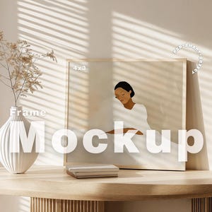 May include: A framed illustration of a person in a white dress sitting on a bed, with the text "4x3" in the top left corner of the frame. The frame is on a light wood table with a white vase and dried flowers.