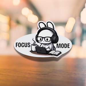 Puede incluir: Una pegatina blanca con un conejo de dibujos animados con auriculares y gafas, trabajando en un portátil con una taza de café y un libro. La pegatina tiene las palabras "FOCUS MODE" en texto negro.