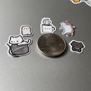 Puede incluir: Una colecci&oacute;n de peque&ntilde;as pegatinas de estilo caricatura con varios dise&ntilde;os lindos. Las pegatinas incluyen un gato blanco sobre una bola de hilo, un fantasma, un cactus en una taza, un gato y un gato marr&oacute;n. Se incluye una moneda como referencia de tama&ntilde;o.