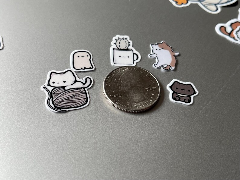 Puede incluir: Una colecci&oacute;n de peque&ntilde;as pegatinas de estilo caricatura con varios dise&ntilde;os lindos. Las pegatinas incluyen un gato blanco sobre una bola de hilo, un fantasma, un cactus en una taza, un gato y un gato marr&oacute;n. Se incluye una moneda como referencia de tama&ntilde;o.