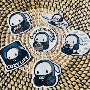Puede incluir: Una colección de pegatinas de conejos blancos con auriculares, cada una con un diseño único. Una pegatina dice "Cozy Life", otra "Overstimulated", y otras presentan frases como "Focus Mode" y "Do Not Disturb". Las pegatinas están dispuestas sobre una cesta tejida.