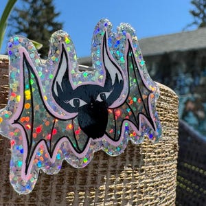 Puede incluir: Pegatina con forma de murciélago brillante con cara negra, orejas blancas y detalles rosas. El murciélago tiene ojos grandes y está cubierto de purpurina iridiscente. Para decoración o manualidades.