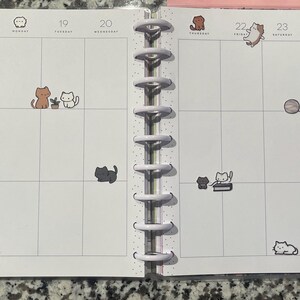 Puede incluir: Una p&aacute;gina de planificador para noviembre con pegatinas con tem&aacute;tica de gatos. Las pegatinas representan varios gatos en diferentes poses, incluyendo sentados, jugando con una pelota y leyendo. El planificador tiene una encuadernaci&oacute;n en espiral y un dise&ntilde;o de cuadr&iacute;cula.