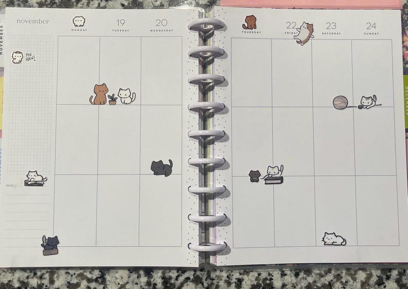 Puede incluir: Una p&aacute;gina de planificador para noviembre con pegatinas con tem&aacute;tica de gatos. Las pegatinas representan varios gatos en diferentes poses, incluyendo sentados, jugando con una pelota y leyendo. El planificador tiene una encuadernaci&oacute;n en espiral y un dise&ntilde;o de cuadr&iacute;cula.