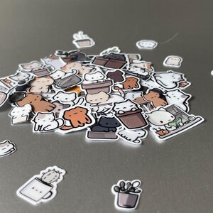 Puede incluir: Una colecci&oacute;n de pegatinas con tem&aacute;tica de gatos en varias poses y colores, incluyendo blanco, marr&oacute;n y negro. Algunas pegatinas presentan gatos en macetas, tazas y otros objetos. Las pegatinas est&aacute;n esparcidas sobre una superficie gris.