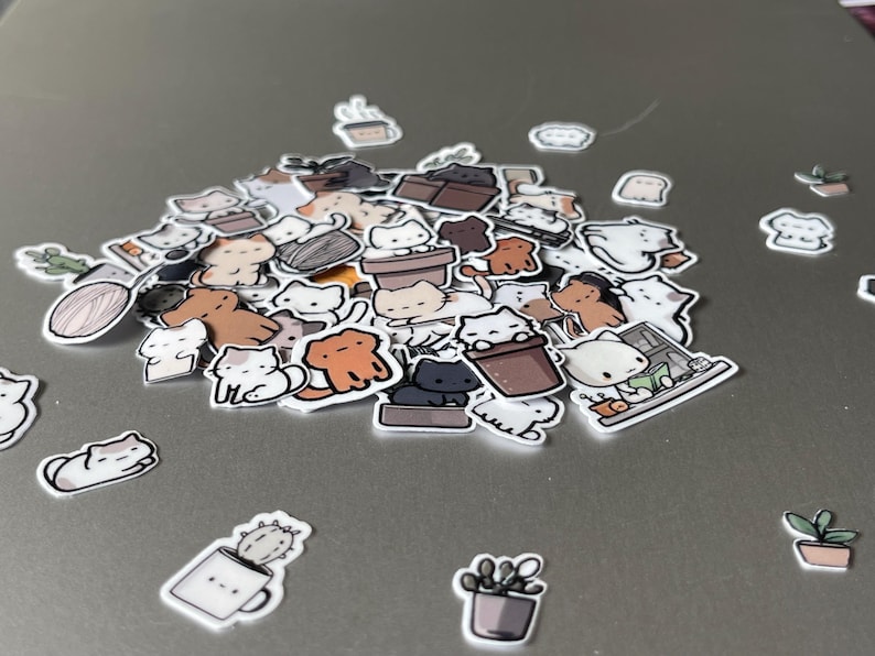Puede incluir: Una colecci&oacute;n de pegatinas con tem&aacute;tica de gatos en varias poses y colores, incluyendo blanco, marr&oacute;n y negro. Algunas pegatinas presentan gatos en macetas, tazas y otros objetos. Las pegatinas est&aacute;n esparcidas sobre una superficie gris.