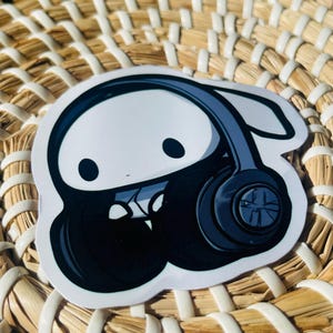 Puede incluir: Una pegatina blanca con un personaje de dibujos animados con auriculares negros y grises. La pegatina está sobre una superficie beige tejida.