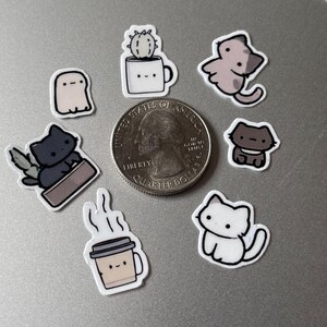 Puede incluir: Una colecci&oacute;n de peque&ntilde;as pegatinas coloridas con varias ilustraciones lindas. Los dise&ntilde;os incluyen un fantasma, gatos, un cactus en una taza y una taza de caf&eacute;. Se incluye una moneda de veinticinco centavos de d&oacute;lar estadounidense como referencia de tama&ntilde;o. Las pegatinas probablemente sean para decorar art&iacute;culos.