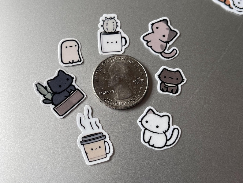Puede incluir: Una colecci&oacute;n de peque&ntilde;as pegatinas coloridas con varias ilustraciones lindas. Los dise&ntilde;os incluyen un fantasma, gatos, un cactus en una taza y una taza de caf&eacute;. Se incluye una moneda de veinticinco centavos de d&oacute;lar estadounidense como referencia de tama&ntilde;o. Las pegatinas probablemente sean para decorar art&iacute;culos.