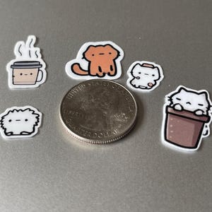 Puede incluir: Una colecci&oacute;n de peque&ntilde;as pegatinas coloridas con gatos de dibujos animados y una taza de caf&eacute;. Las pegatinas est&aacute;n en varias poses y dise&ntilde;os, con una moneda de veinticinco centavos para comparar el tama&ntilde;o. Las pegatinas son blancas con contornos negros.