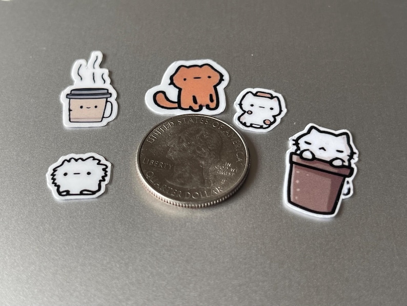 Puede incluir: Una colecci&oacute;n de peque&ntilde;as pegatinas coloridas con gatos de dibujos animados y una taza de caf&eacute;. Las pegatinas est&aacute;n en varias poses y dise&ntilde;os, con una moneda de veinticinco centavos para comparar el tama&ntilde;o. Las pegatinas son blancas con contornos negros.