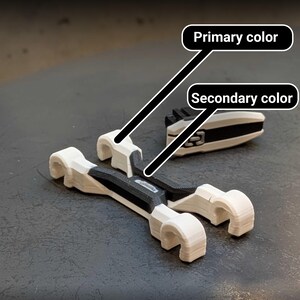 May include: Two white and black 3D-printed objects are displayed on a dark surface. One object is a pair of curved, wrench-like shapes. The other is a rectangular piece with a black center and white edges. The objects appear to be components of a larger mechanism.