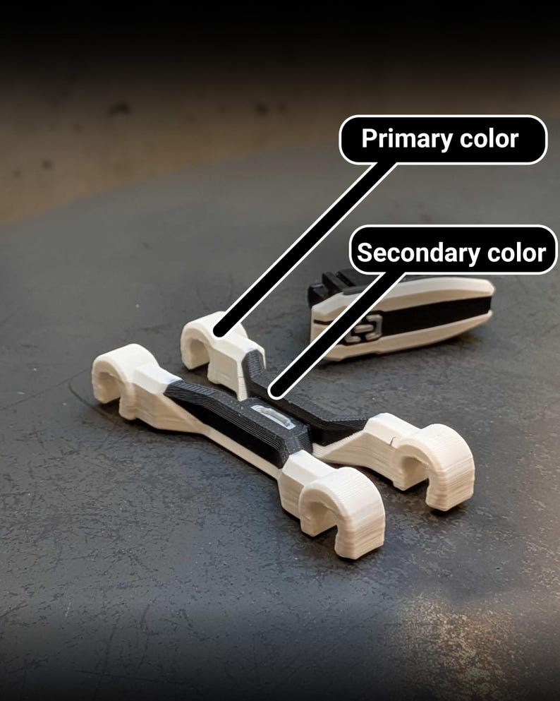 May include: Two white and black 3D-printed objects are displayed on a dark surface. One object is a pair of curved, wrench-like shapes. The other is a rectangular piece with a black center and white edges. The objects appear to be components of a larger mechanism.