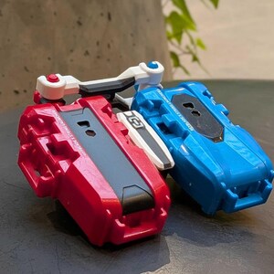 May include: Two toy launchers, one red and one blue, are shown. The red launcher has a black center and white accents. The blue launcher has a black center and white accents. Both have a handle and are designed for launching toys.