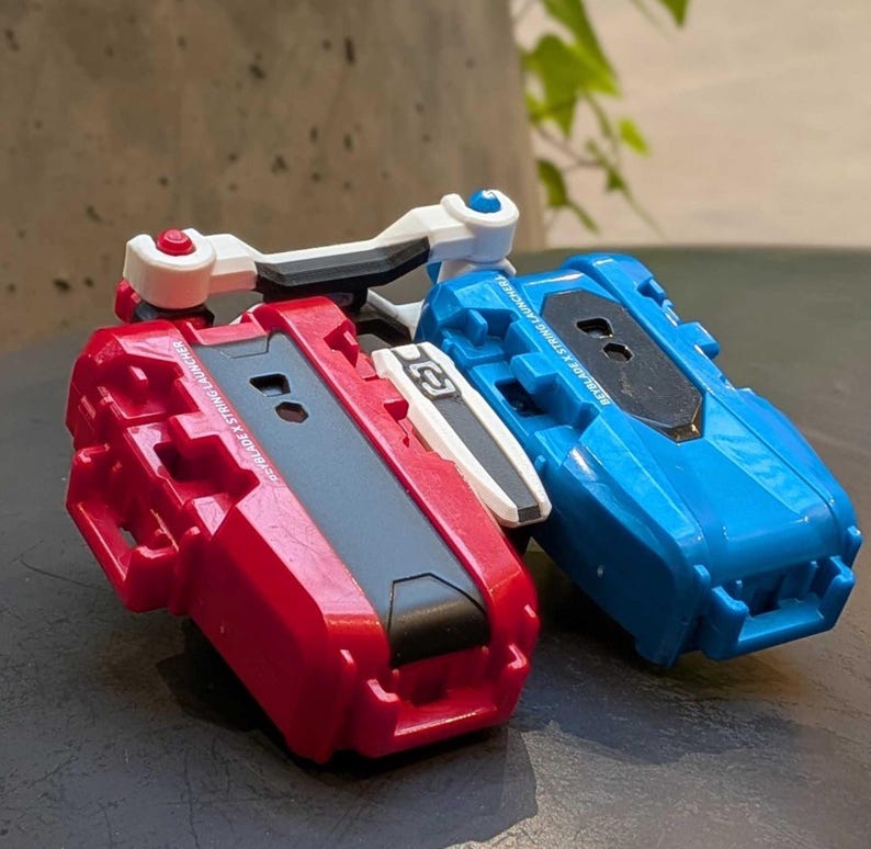 May include: Two toy launchers, one red and one blue, are shown. The red launcher has a black center and white accents. The blue launcher has a black center and white accents. Both have a handle and are designed for launching toys.