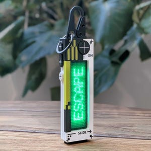Puede incluir: Un llavero electrónico rectangular blanco y negro con una pantalla LED verde brillante que muestra la palabra ESCAPE. El dispositivo incluye un mosquetón negro, un botón circular de encendido ON, un botón de signo más y el texto SLIDE con una flecha.