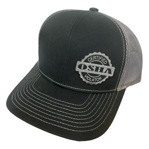 Könnte beinhalten: Eine schwarze und graue Trucker-Kappe mit einem silbernen Patch, auf dem "Certified OSHA Violator" steht.