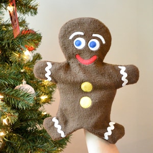 May include: A brown gingerbread man puppet with white icing, yellow buttons, and a red smiling mouth. The puppet has blue eyes and is being held up by a hand.