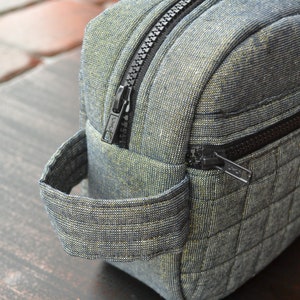 May include: A gray quilted toiletry bag with a black zipper and a black handle. The bag has a small zippered pocket on the side.