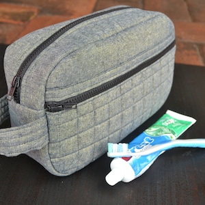 May include: A gray quilted toiletry bag with a black zipper and a handle. The bag is sitting on a black surface with a tube of toothpaste and a toothbrush.