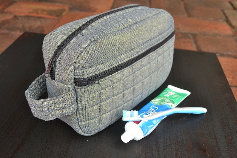 May include: A gray quilted toiletry bag with a black zipper and a handle. The bag is sitting on a black surface with a tube of toothpaste and a toothbrush.