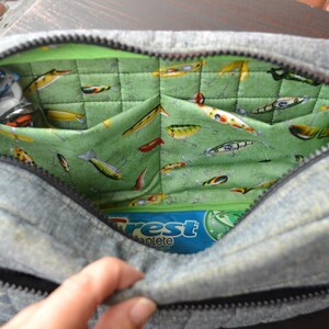 May include: A gray denim zippered pouch with a green quilted lining featuring a pattern of colorful fishing lures. The pouch is open and a blue and white package with the word "Crest" is visible inside.