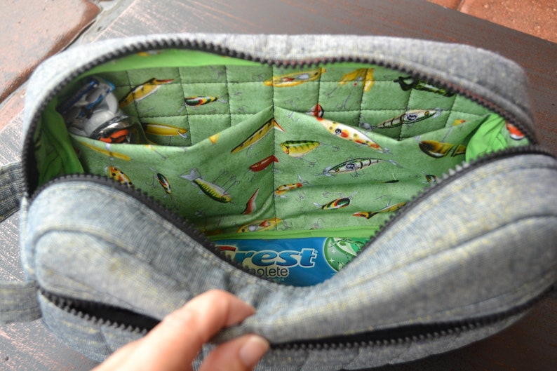 May include: A gray denim zippered pouch with a green quilted lining featuring a pattern of colorful fishing lures. The pouch is open and a blue and white package with the word "Crest" is visible inside.