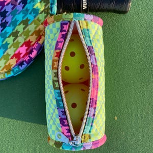 May include: A colorful pickleball set featuring a paddle and a ball holder. The holder is zippered and contains two yellow pickleballs. The paddle handle is black with the text "VUFOX1". The set has a houndstooth pattern in multiple colors.