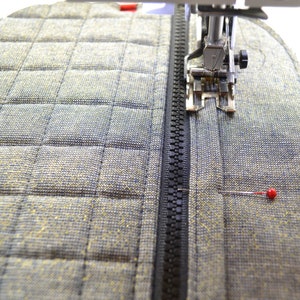 May include: A close-up of a sewing machine stitching a black zipper onto a gray quilted fabric. A red pin is visible in the fabric.