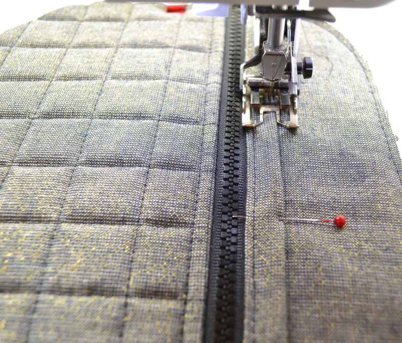 May include: A close-up of a sewing machine stitching a black zipper onto a gray quilted fabric. A red pin is visible in the fabric.