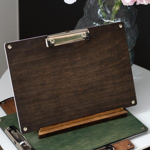 May include: Dark brown wooden clipboard with a silver metal clip, resting on a wooden stand. The clipboard is on top of a green clipboard and a brown clipboard. The background includes a vase with white and pink flowers.
