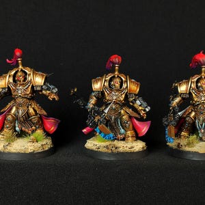 Peut inclure: Trois figurines miniatures peintes en or et noir de guerriers portant des capes rouges et dorées. Les figurines tiennent des armes et sont en position de combat. Les figurines sont debout sur des socles circulaires.