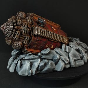 Puede incluir: Una miniatura detallada pintada a mano de un taladro mecánico futurista, descansando sobre un lecho de rocas grises. El taladro está hecho de metal y tiene un esquema de color rojo y marrón. El taladro está cubierto de detalles intrincados, incluyendo engranajes, tuberías y cables.