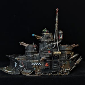 Peut inclure: Maquette détaillée d'un char de guerre noir et bronze avec plusieurs tourelles, une grande lame et divers composants mécaniques. Le modèle présente un personnage Ork et est posé sur un fond noir.