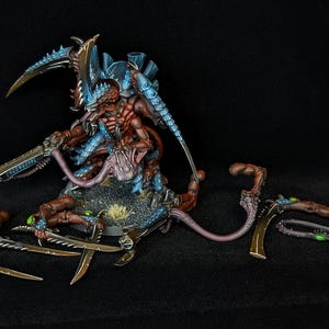 Puede incluir: Figura en miniatura detallada de una criatura con exoesqueleto marrón y azul, múltiples extremidades y cuchillas afiladas. La figura está posada sobre una base texturizada con componentes de armas desmontados adicionales. Tema de ciencia ficción o fantasía.