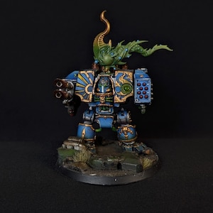 Può includere: Figura in miniatura dettagliata di un robot corazzato blu e oro con un copricapo verde dall'aspetto organico. Il robot ha due grandi canne di fucile e si trova su una base strutturata. La figura è dipinta con dettagli intricati.